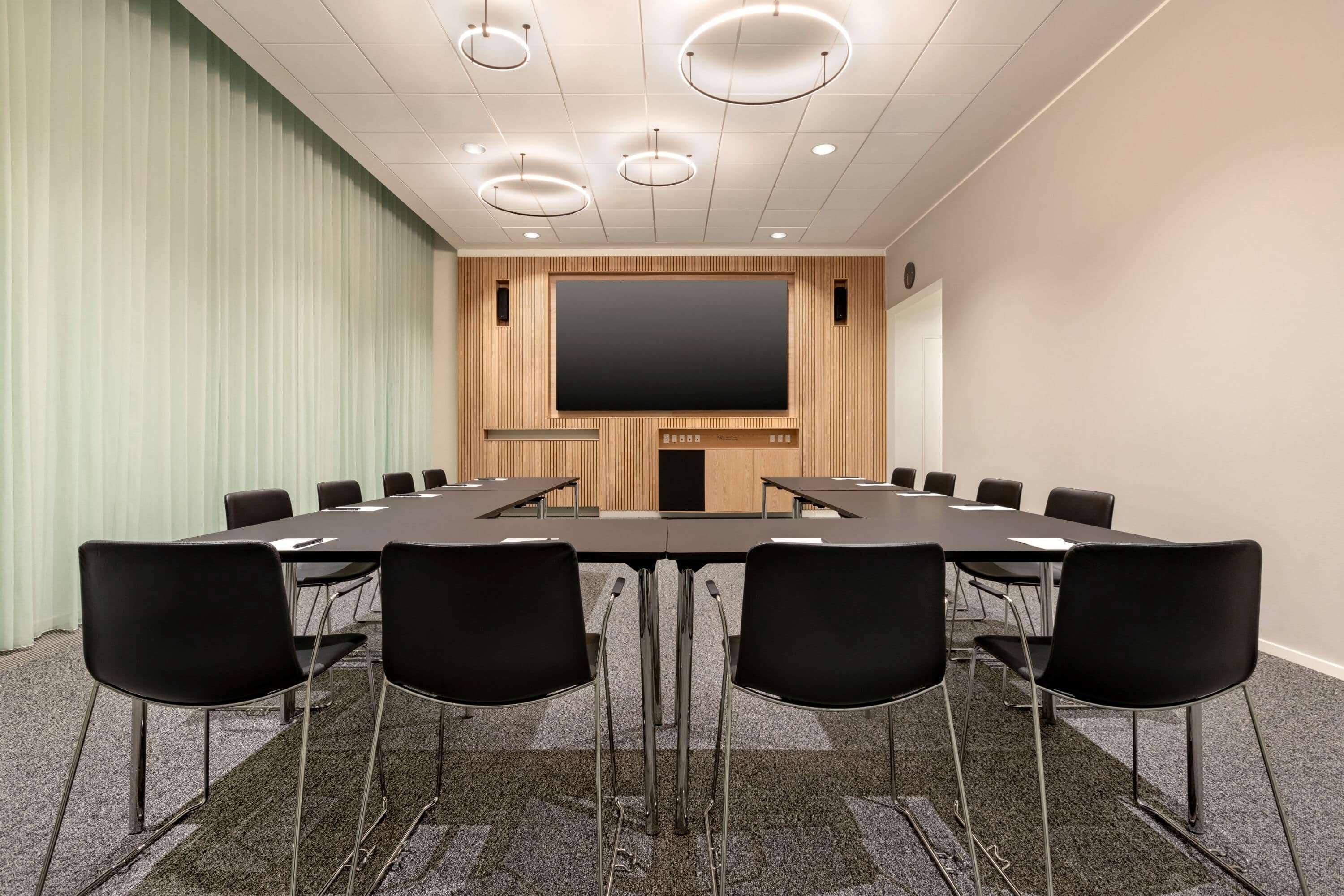 A conference room with a long table, black chairs, a large screen, and a wooden panel wall.