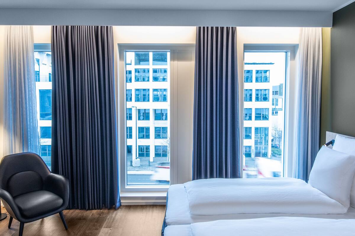 Quarto de hotel com uma cama grande, uma cadeira e uma janela com cortinas.