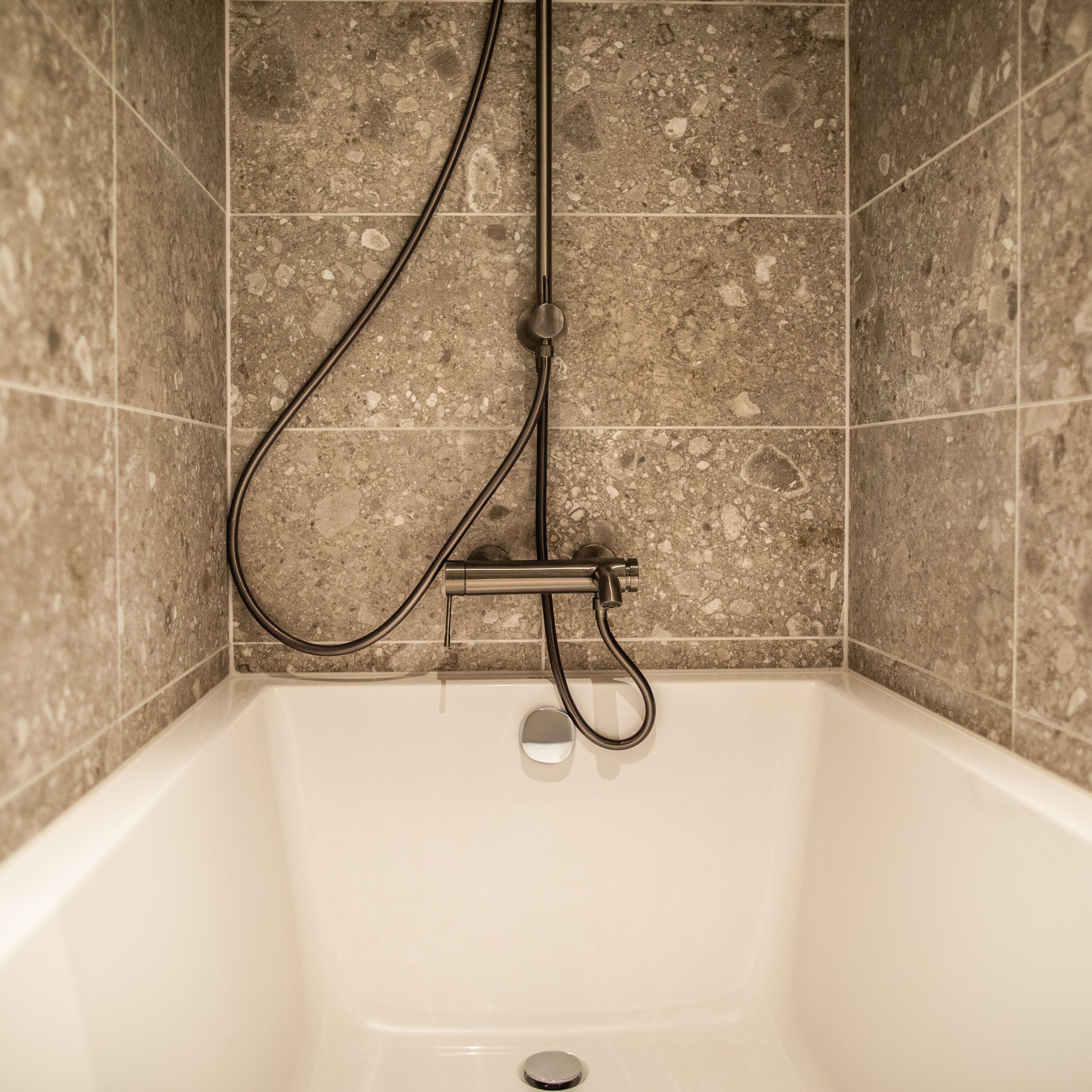 A corner bathtub with a shower hose and controls, set against a speckled tile wall.