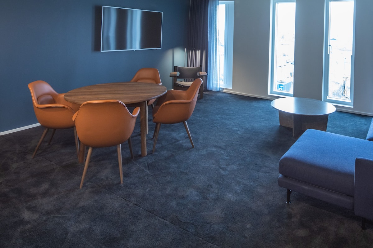 Uma sala de reuniões moderna com uma grande mesa, seis cadeiras, uma TV de tela plana e janelas amplas.