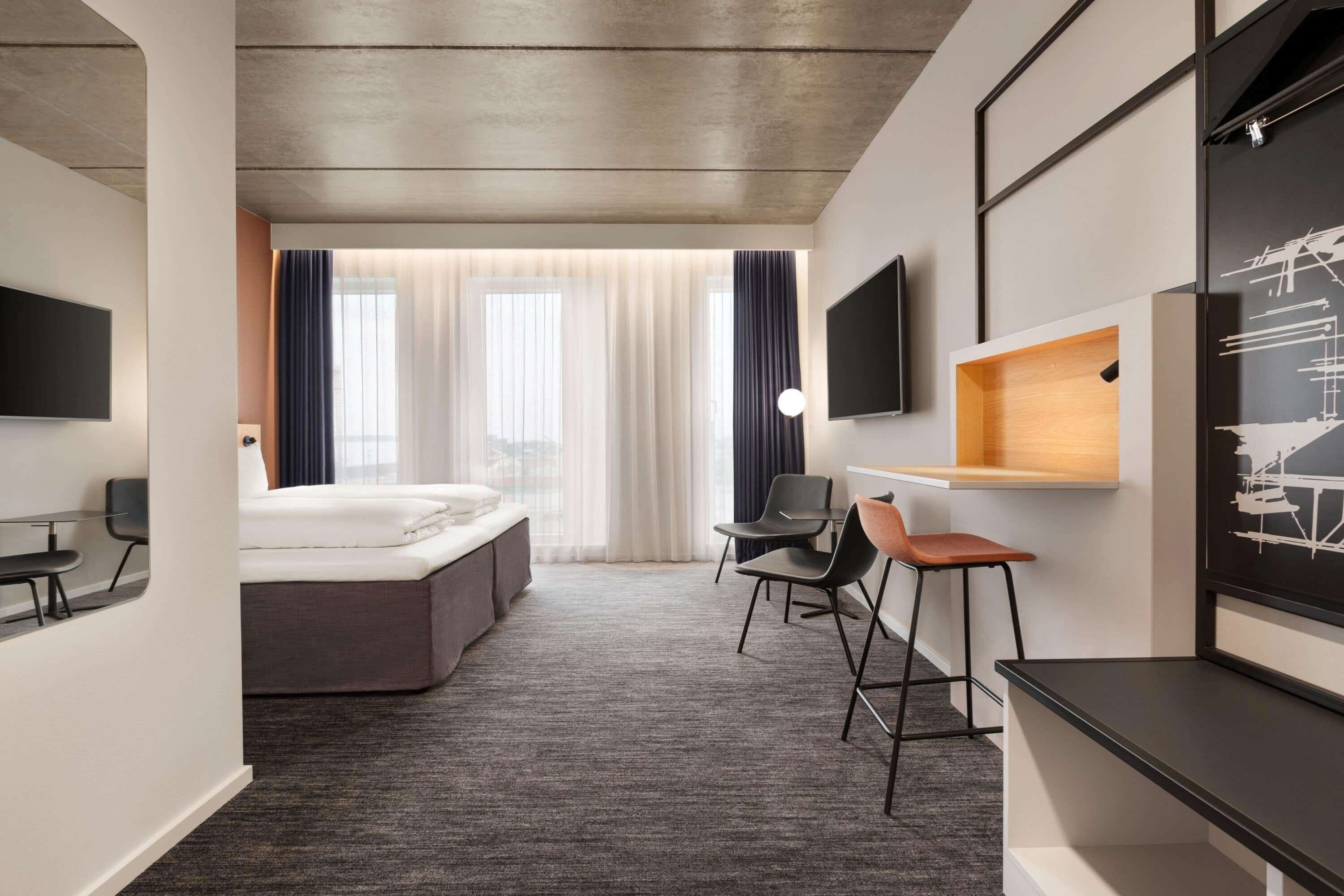 A modern hotel room with a large bed, a desk with a chair, a TV mounted on the wall, and a window with curtains.