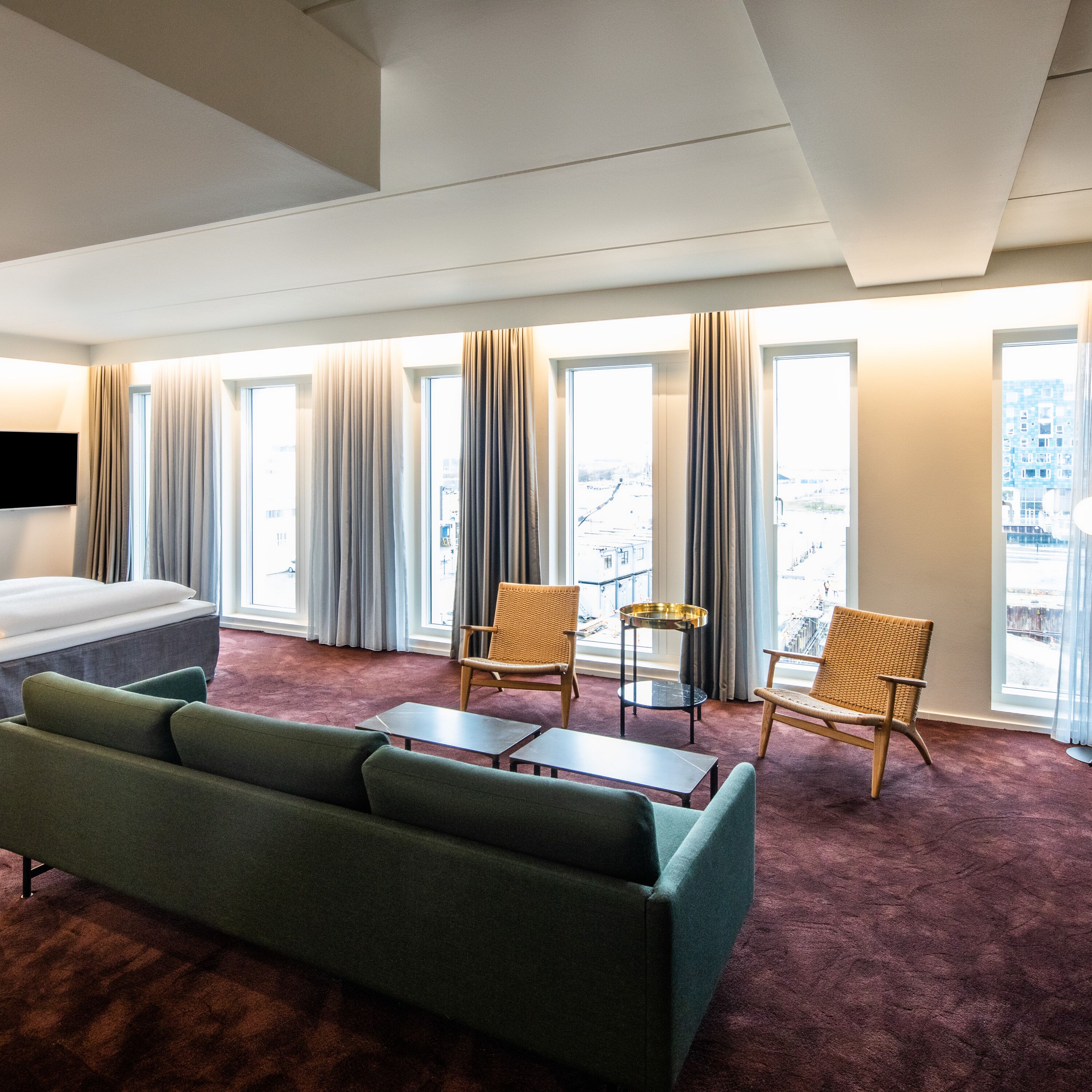 A hotel room with a bed, green sofa, two wicker chairs, a small table, and large windows with curtains.