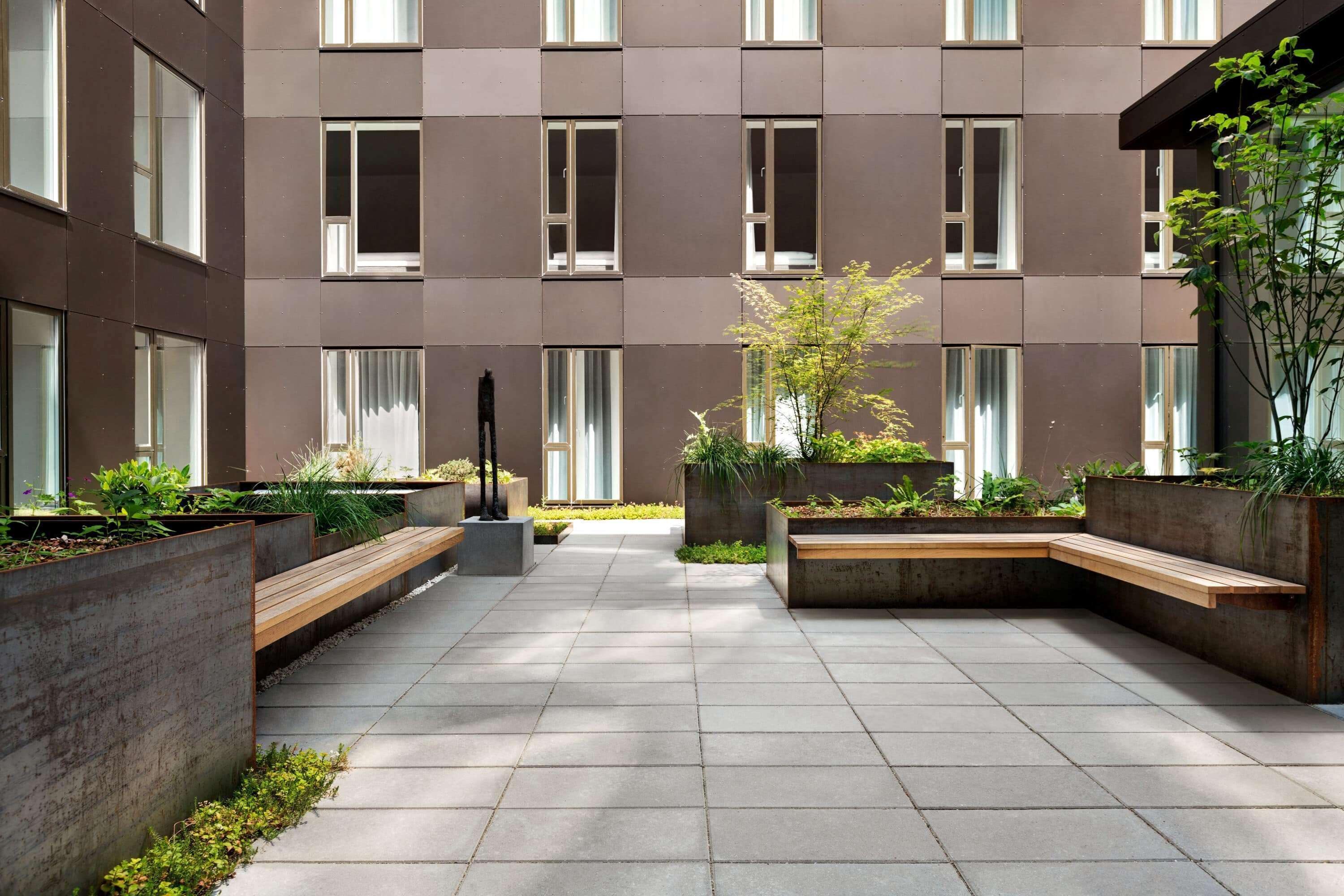 A courtyard with a modern building, concrete planters with greenery, and a central seating area.