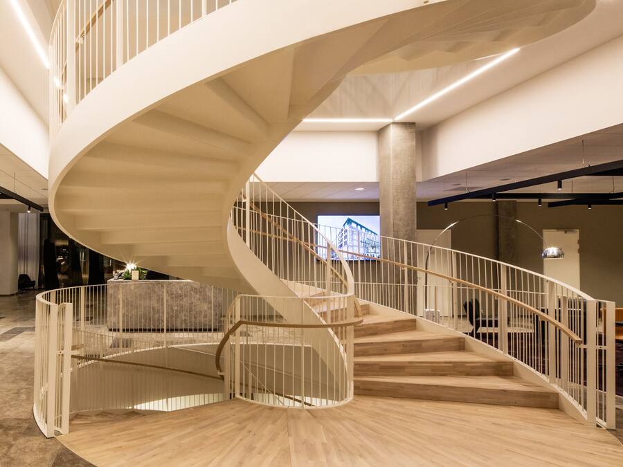 A modern spiral staircase with a wooden finish and white railings, surrounded by a spacious, open-plan area with a large screen displaying a cityscape.