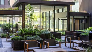 An outdoor seating area with wooden furniture and planters, surrounded by glass walls and a modern building.