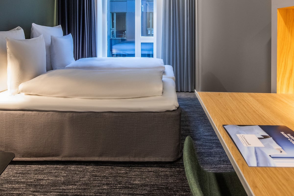Quarto de hotel com cama, escrivaninha, televisão e vista do interior através de uma porta.