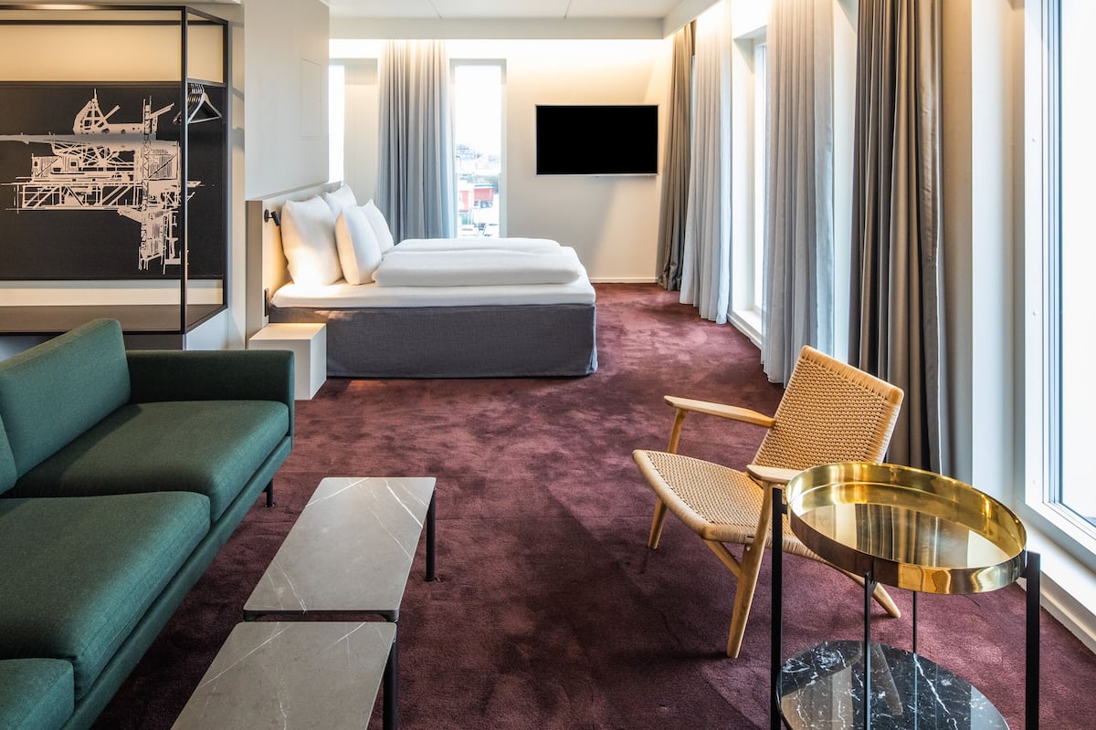 Quarto de hotel com cama, sofá verde, mesa pequena, cadeira de vime e uma grande janela com cortinas.