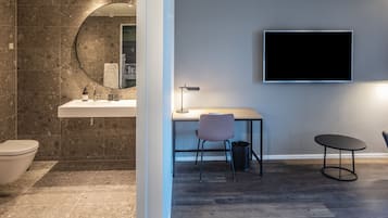 Et moderne badeværelse med vask, spejl og toilet. Der er et TV ophængt på væggen og et lille bord med en stol.