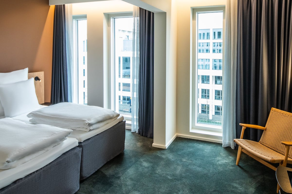 Quarto de hotel com cama, uma cadeira e janelas amplas com vista para a cidade.