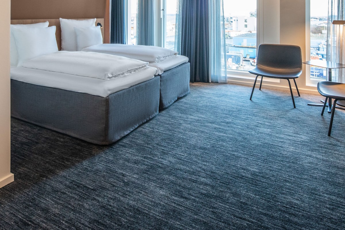 Quarto de hotel com uma cama grande, uma escrivaninha e uma cadeira. Há cortinas e vista para a cidade pelas janelas.