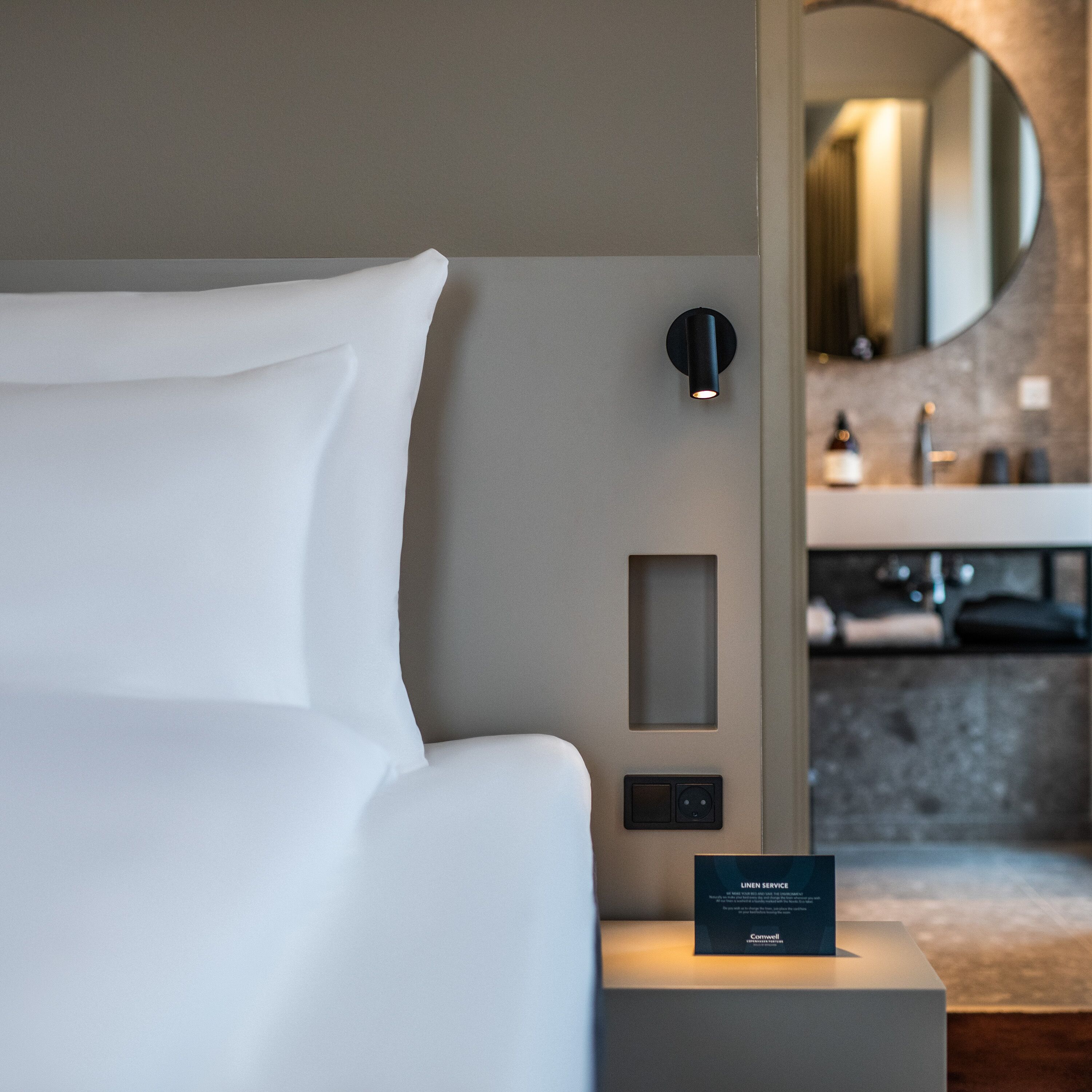 Una habitación de hotel moderna con una cama bien hecha, una lámpara fijada a la pared, un pequeño panel de control y una tarjeta de servicio de ropa.
