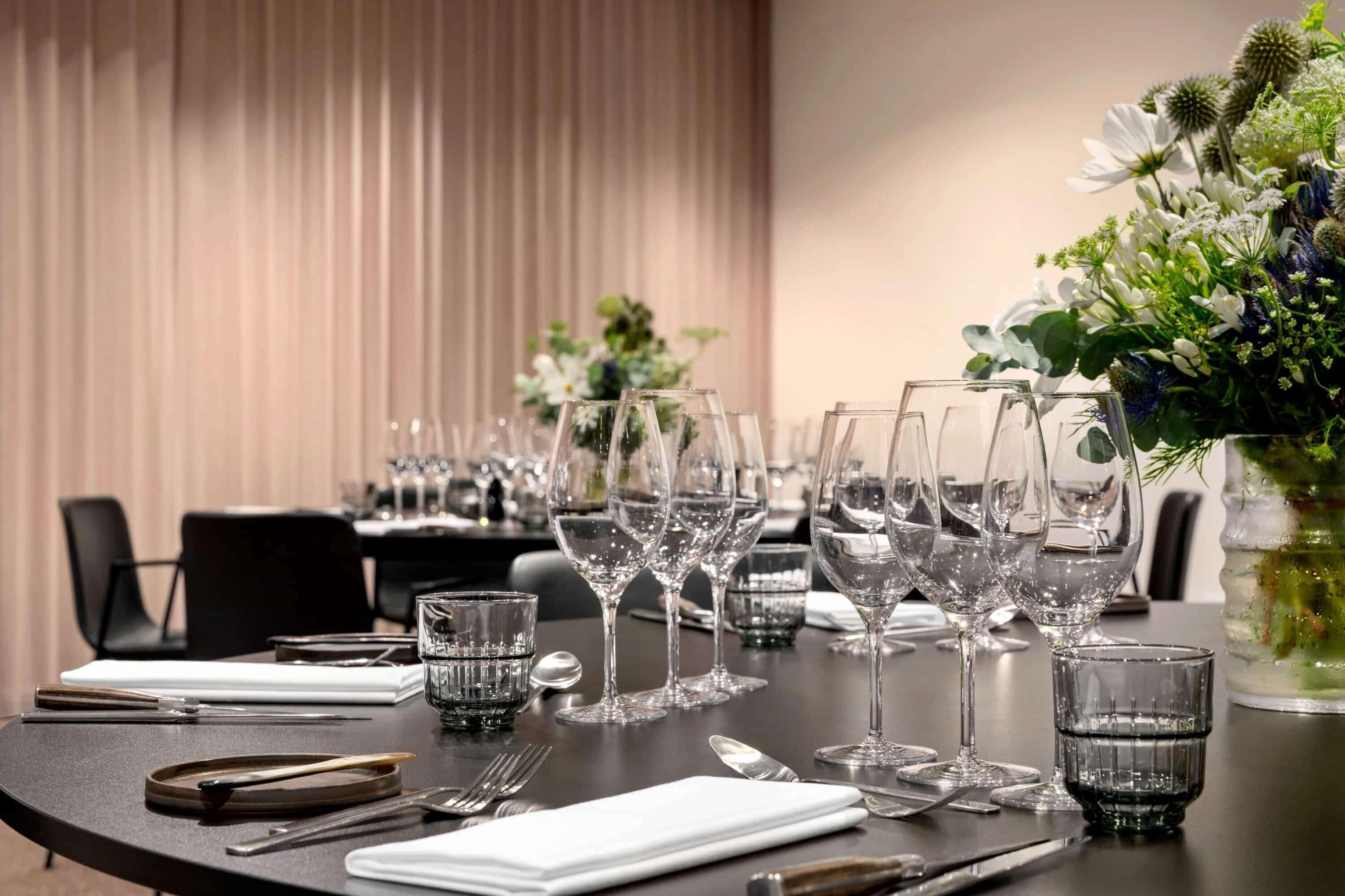 A dining table set with multiple wine glasses, water glasses, and cutlery, with a floral centerpiece and a curtain in the background.