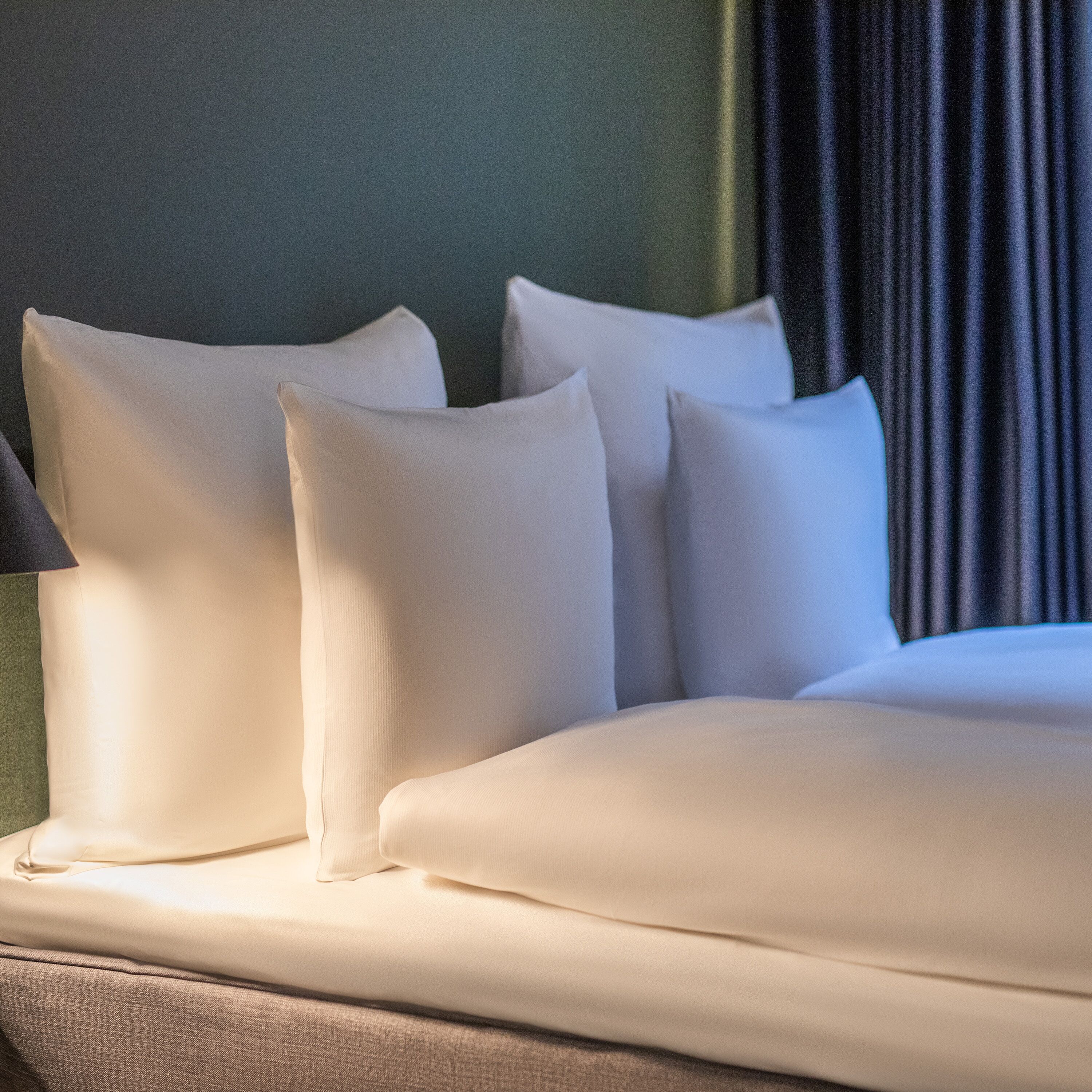 Una cama bien hecha con sábanas y almohadas blancas, una pared verde y una cortina oscura al fondo.