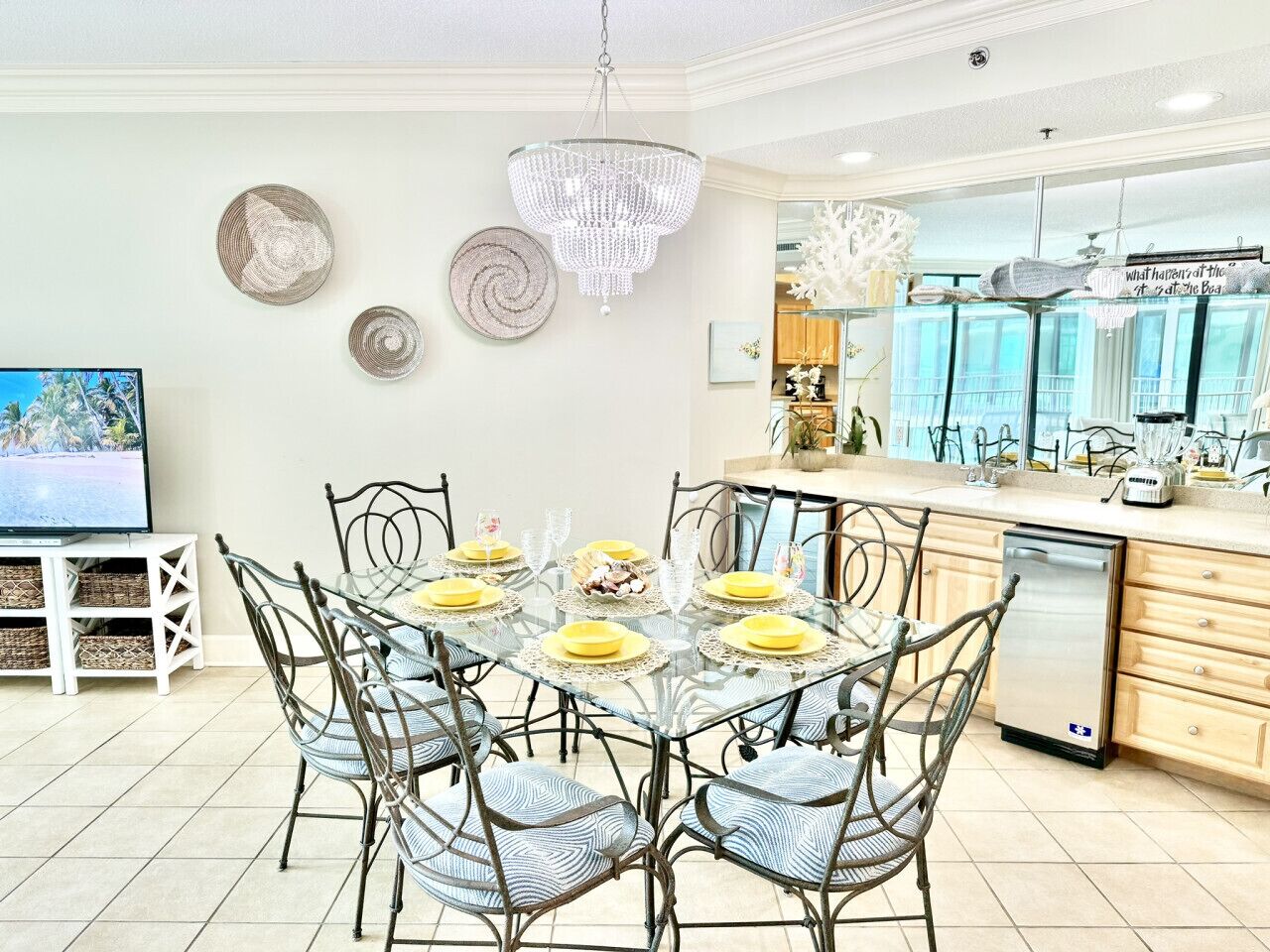 Un coin repas avec une table en verre dressée pour six personnes, entourée de chaises en métal. Un lustre est suspendu au-dessus, et un téléviseur est posé sur un meuble à gauche.