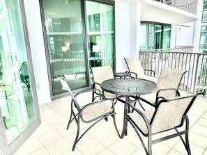 Een balkon met een ronde tafel en vier stoelen, glazen deuren en uitzicht op een gebouw met balkons.