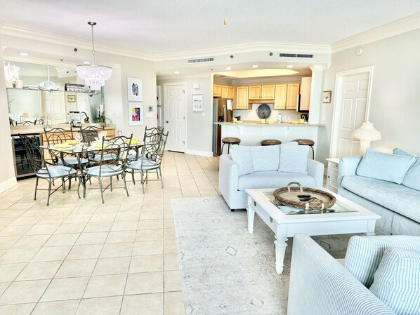 A spacious living room with a dining area, a kitchen in the background, and a large chandelier.