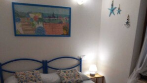 Een bed met een blauw hoofdeinde, een witte dekbedovertrek en twee kussens met een patroon. Daarboven hangt een schilderij aan de muur en ernaast staat een tafeltje met een lamp.