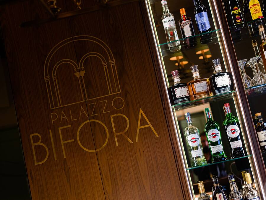 A wooden bar counter with the name "Palazzo Bifora" and a variety of liquor bottles.
