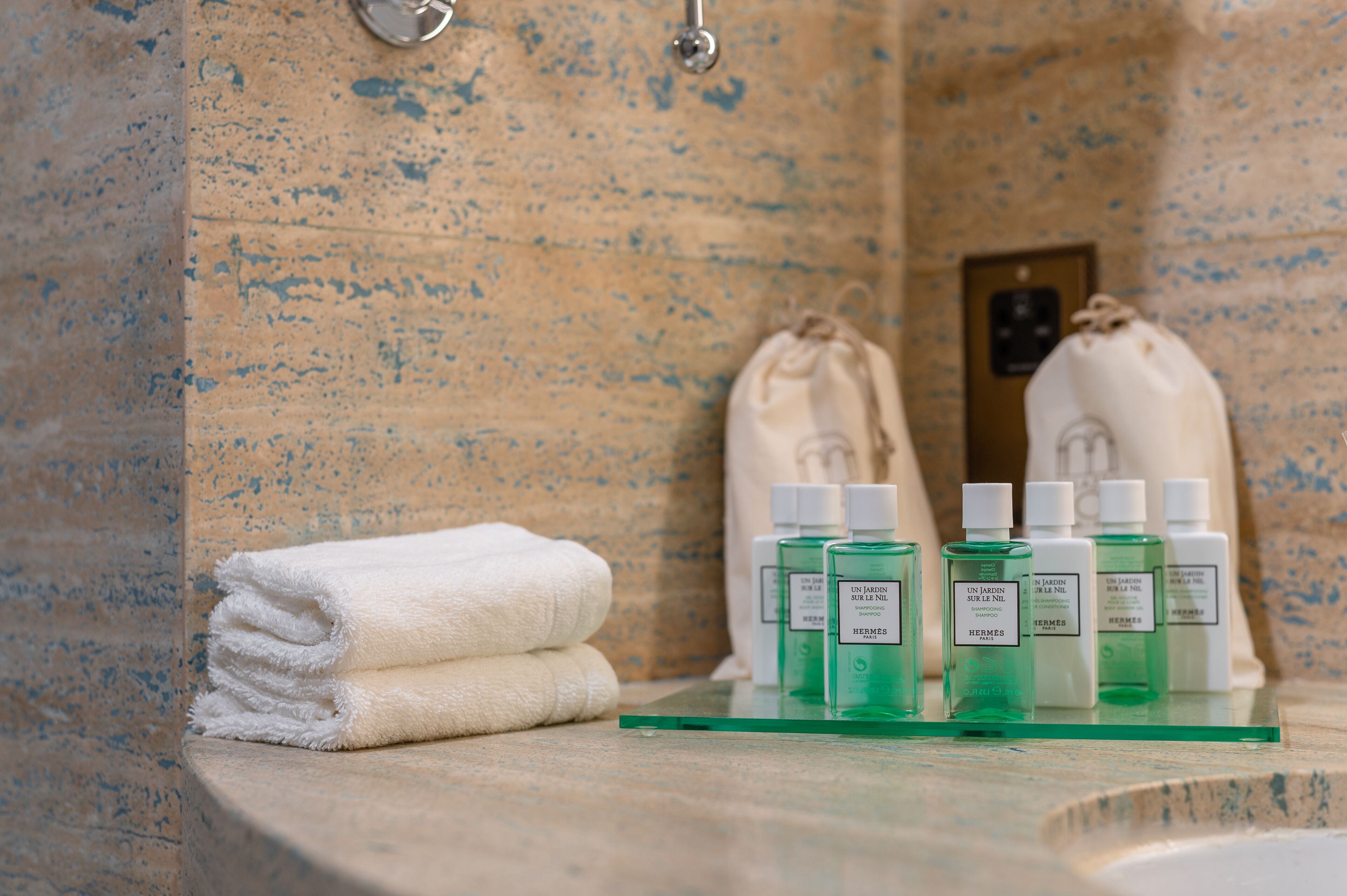 A bathroom with a marble wall, a towel, and several bottles of green liquid on a glass shelf.