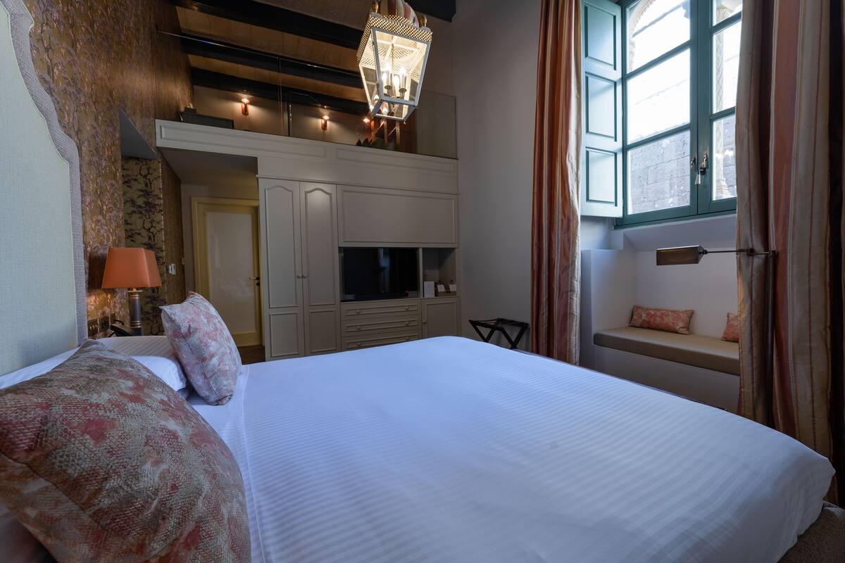 Habitación de hotel con una cama grande, televisión, zona de estar con un banco y ventana con cortinas.