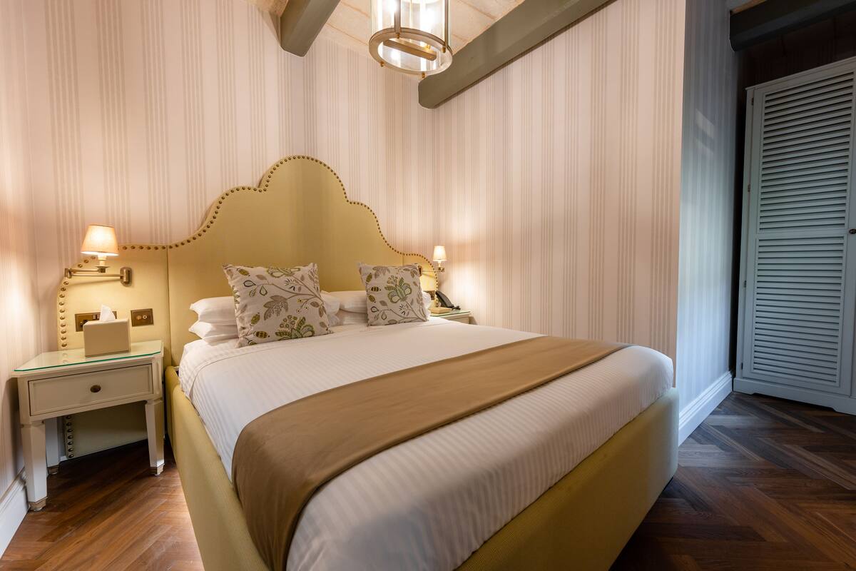 Una cama bien tendida con un cabecero dorado, dos mesitas de noche con lámparas y una puerta blanca al fondo.