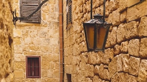 A narrow stone alleyway with a red door, a potted plant, and a lantern. - Palazzo Bifora (Mdina)