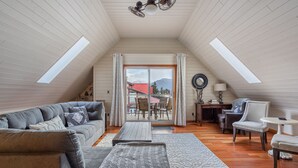 A cozy loft-style living room with a high ceiling, wooden floors, a sectional sofa, a coffee table, and a view of a mountain through a large sliding glass door.