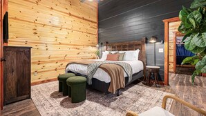 A wooden cabin bedroom with a bed, two green stools, a small table, and a large plant. - Heated In-Cabin Pool~Theatre~Game Room~Views (Sevierville)