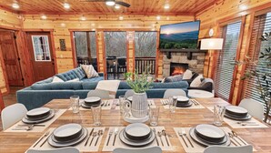 L'intérieur d'un chalet en bois comprend une table à manger pour six personnes, une cheminée et un grand téléviseur fixé au mur.