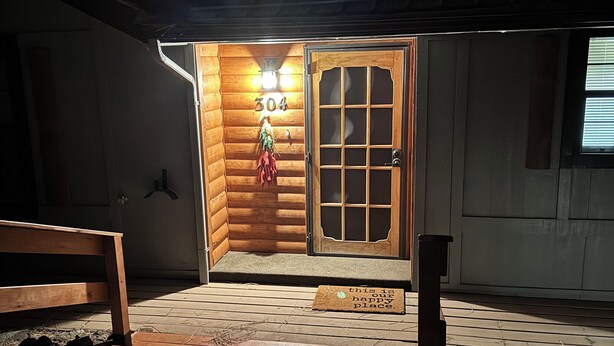 Entrada de una cabaña de madera con un letrero con el número '304', un banco y un felpudo con un mensaje.