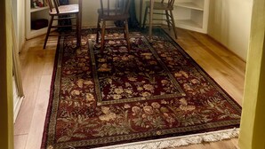 Ein Zimmer mit Holztisch, zwei Stühlen, einem gemusterten Teppich, einem Fenster mit Vorhängen und einer eingebauten Regalanlage mit verschiedenen Gegenständen.