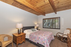 A bedroom with a wooden ceiling, a bed with a floral bedspread, two bedside lamps, a wicker chair, and a window with blinds. - Cozy Escape- Walking Distance to Village! (Lake Arrowhead)