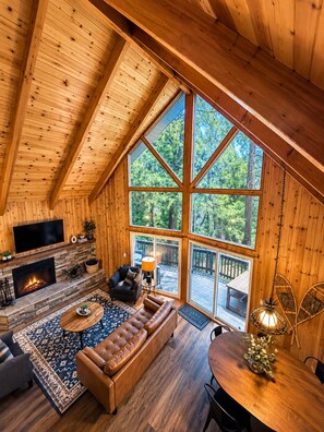A log cabin interior with a fireplace, leather sofa, and a large window overlooking a deck. - Cozy Escape- Walking Distance to Village! (Lake Arrowhead)