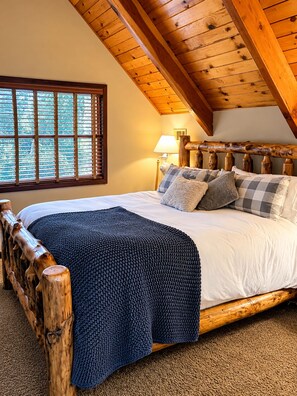 A bedroom with a wooden headboard, a large bed with multiple pillows, a window with blinds, and a lamp on the nightstand.