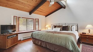 Un dormitorio con techo de madera, una cama con estructura de metal, un televisor sobre un mueble de madera y una ventana con persianas.
