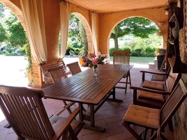Un porche cubierto con una mesa y sillas de madera, rodeado de arcos de ladrillo y plantas en macetas.