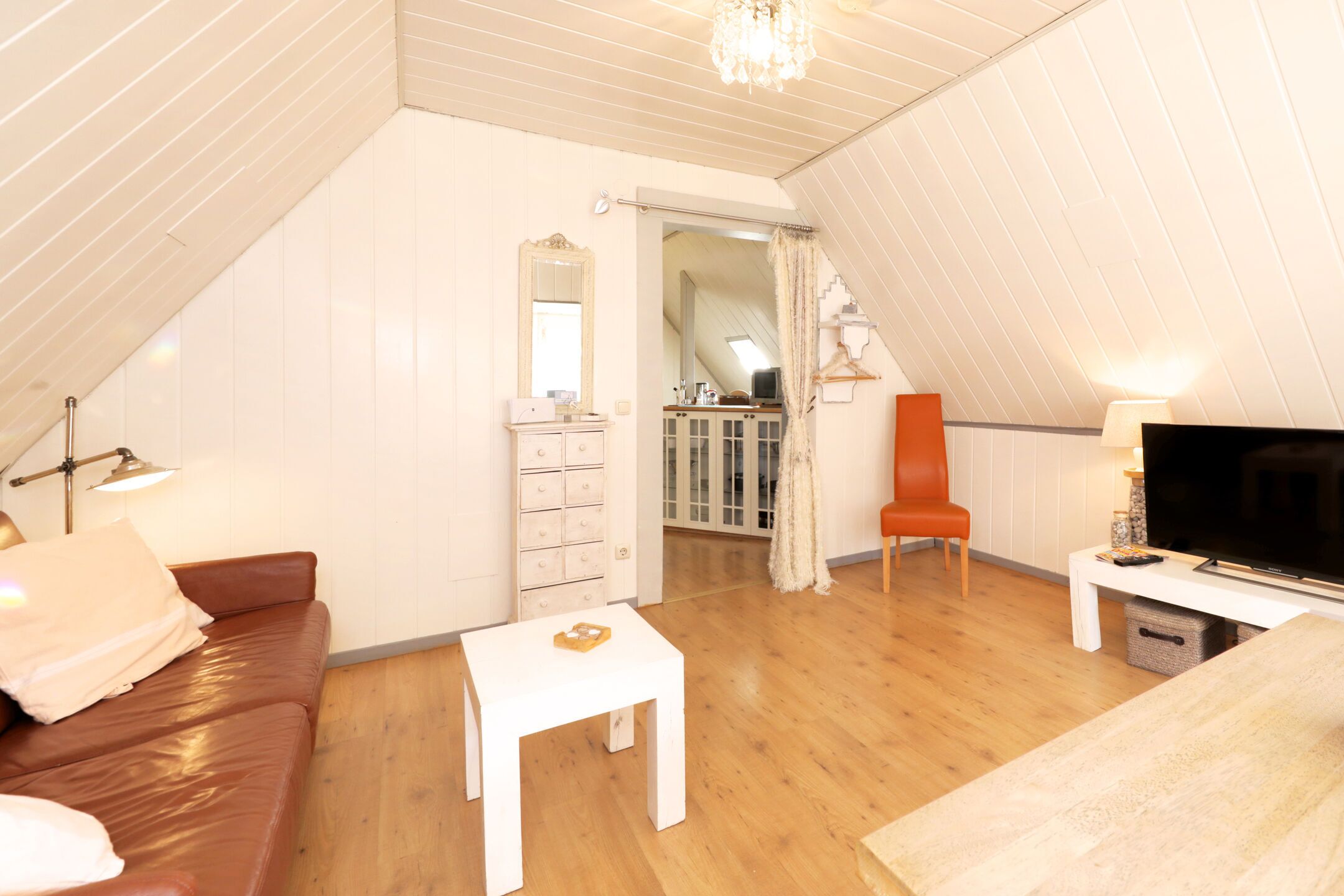 Une chambre confortable située dans les combles, avec un canapé en cuir marron, une table blanche, une télévision et un escalier menant à une salle de bain.