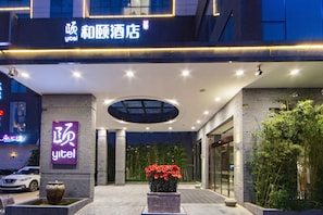A hotel entrance with a modern design, featuring a large illuminated sign, a central flower arrangement, and a well-lit facade.