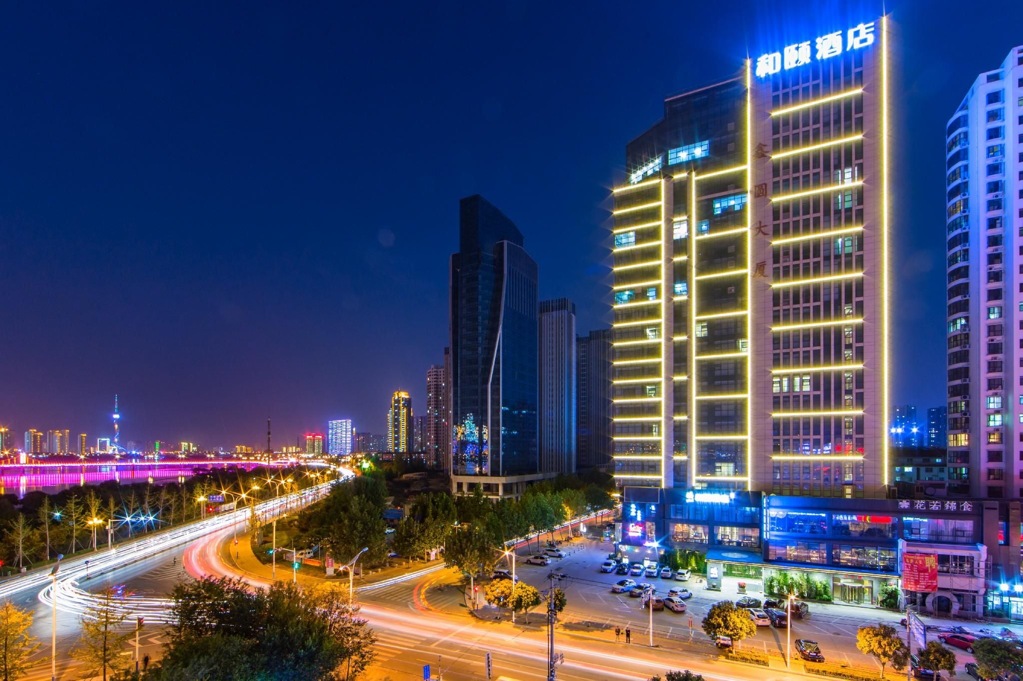 A multi-story hotel with illuminated signage, a parking area, and a busy road with light trails.