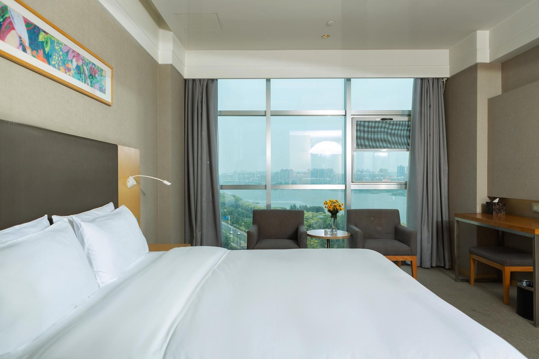 Een hotelkamer met een groot bed, twee fauteuils, een tafeltje en uitzicht op een stadsgezicht door grote ramen.