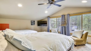 A bedroom with two beds, a ceiling fan, a TV, and a large window with a view of trees and a road.