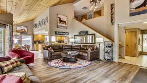 A spacious living area with a leather sectional sofa, a round coffee table, and a colorful rug. A kitchen with wooden cabinets and a farmhouse sign is visible in the background. - Conifer 10 (Bend)