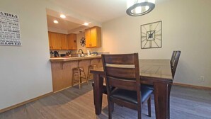 A dining area with a wooden table and chairs, a kitchen in the background, and a wall with a clock and a poster. - Skyline Condo 02 (Bend)