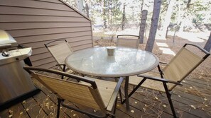 A round metal table with four wicker chairs, a grill, and a metal trash can on a wooden deck.