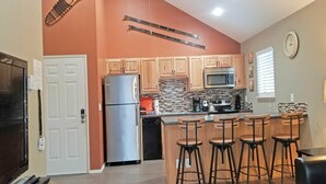 A kitchen with wooden cabinets, a stainless steel refrigerator, a microwave, and a coffee maker. There are four bar stools and a window with blinds. - Powder Village E2 (Bend)