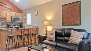 A living room with a black leather sofa, a coffee table, and a kitchen area with wooden cabinets and a microwave. - Powder Village E2 (Bend)
