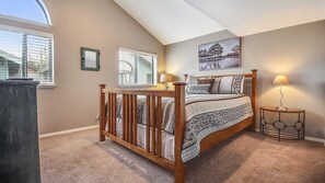 A bedroom with a sloped ceiling, a wooden crib, a bed with patterned bedding, two bedside lamps, and a framed picture on the wall. - Powder Village E2 (Bend)