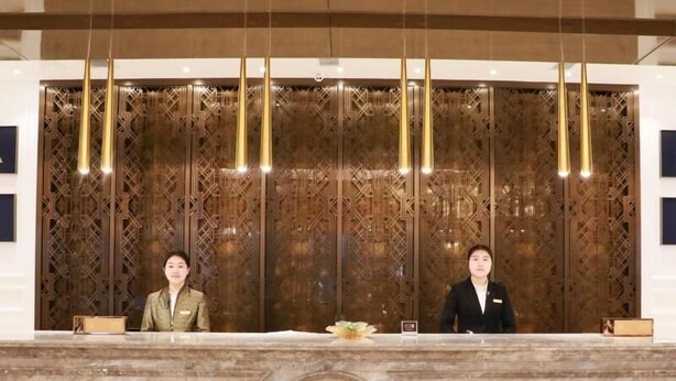 A reception desk with two staff members, a marble counter, and a decorative wooden panel with hanging lights. - Greentree Eastern Yancheng Binhai County Aobaolia Beichen Hotel (Yancheng)