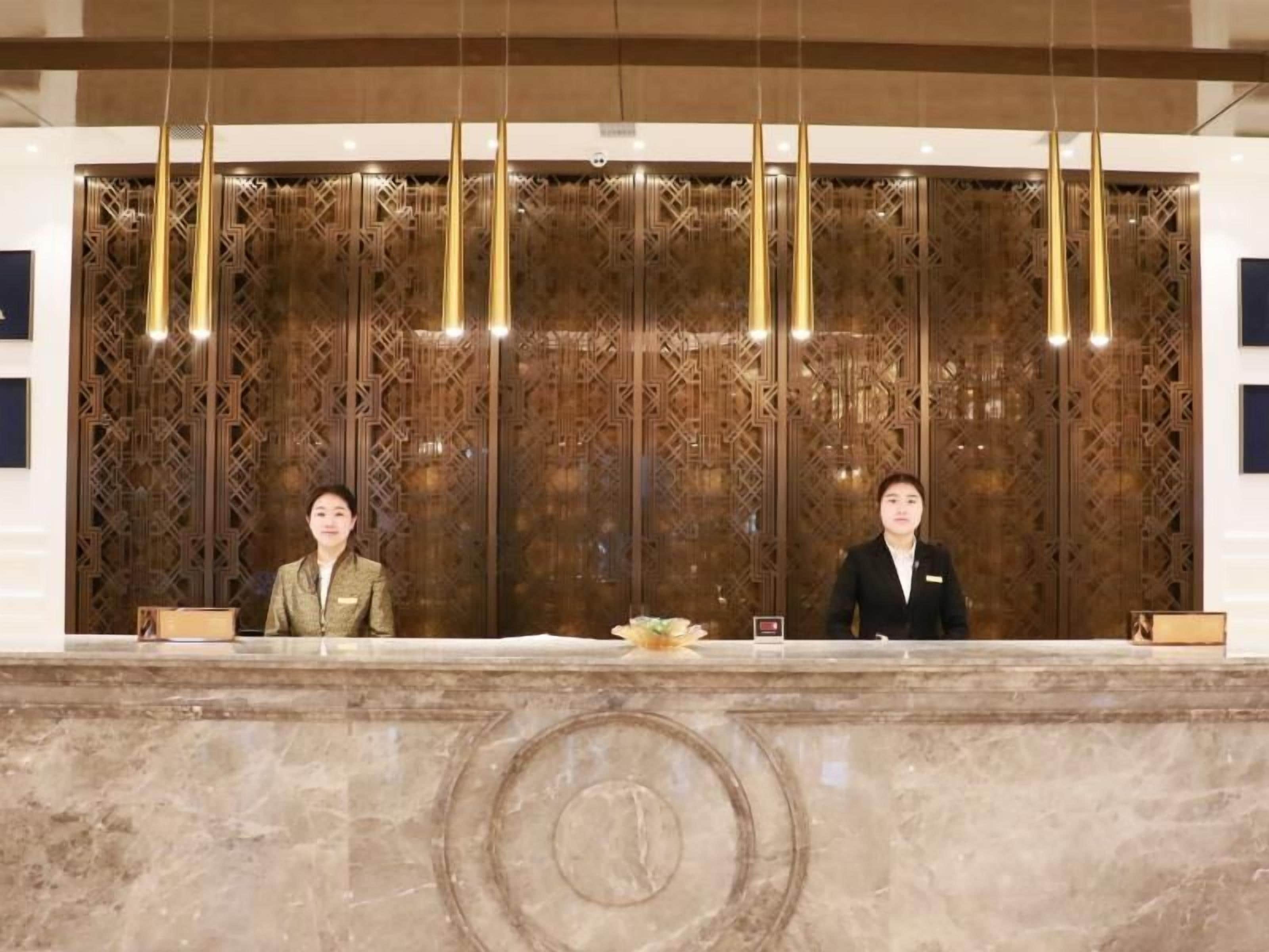 Una recepción con dos empleados, un mostrador de mármol y un panel de madera decorativo con luces colgantes.