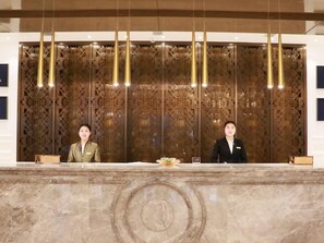 A reception desk with two staff members, a marble counter, and a decorative wooden panel with hanging lights.